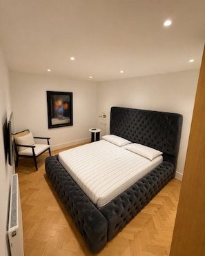 Quarto com uma cama grande, uma cadeira, uma mesinha e uma televisão fixada na parede.