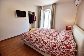 A bedroom with a bed, a TV mounted on the wall, a window with curtains, and an air conditioning unit.