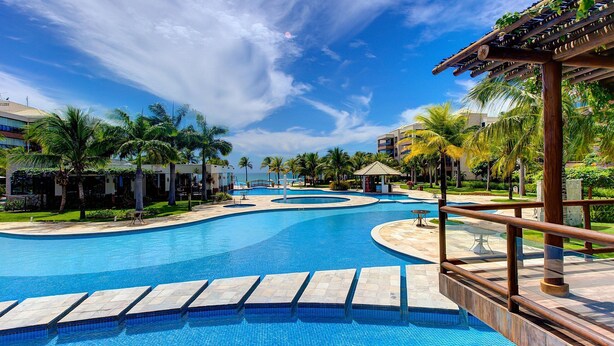 A large, clear swimming pool with a curved design, surrounded by palm trees and a wooden pergola. - Waiwai Cumbuco pé na areia por Carpediem (Caucaia)