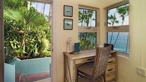 Een kamer met een houten bureau, een rieten stoel, uitzicht op de oceaan en tropische planten.
