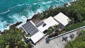 Um edifício costeiro com painéis solares, rodeado por palmeiras e terreno rochoso.