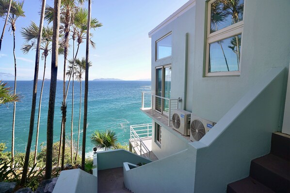 Un bâtiment moderne doté d’un balcon donnant sur une mer d’un bleu éclatant, entouré de palmiers.