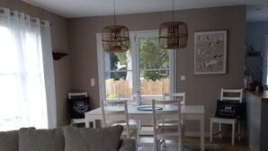 Een woonkamer met een grijze bank, een eettafel met stoelen, twee hangende rieten lampen en een raam met uitzicht op een houten schutting.