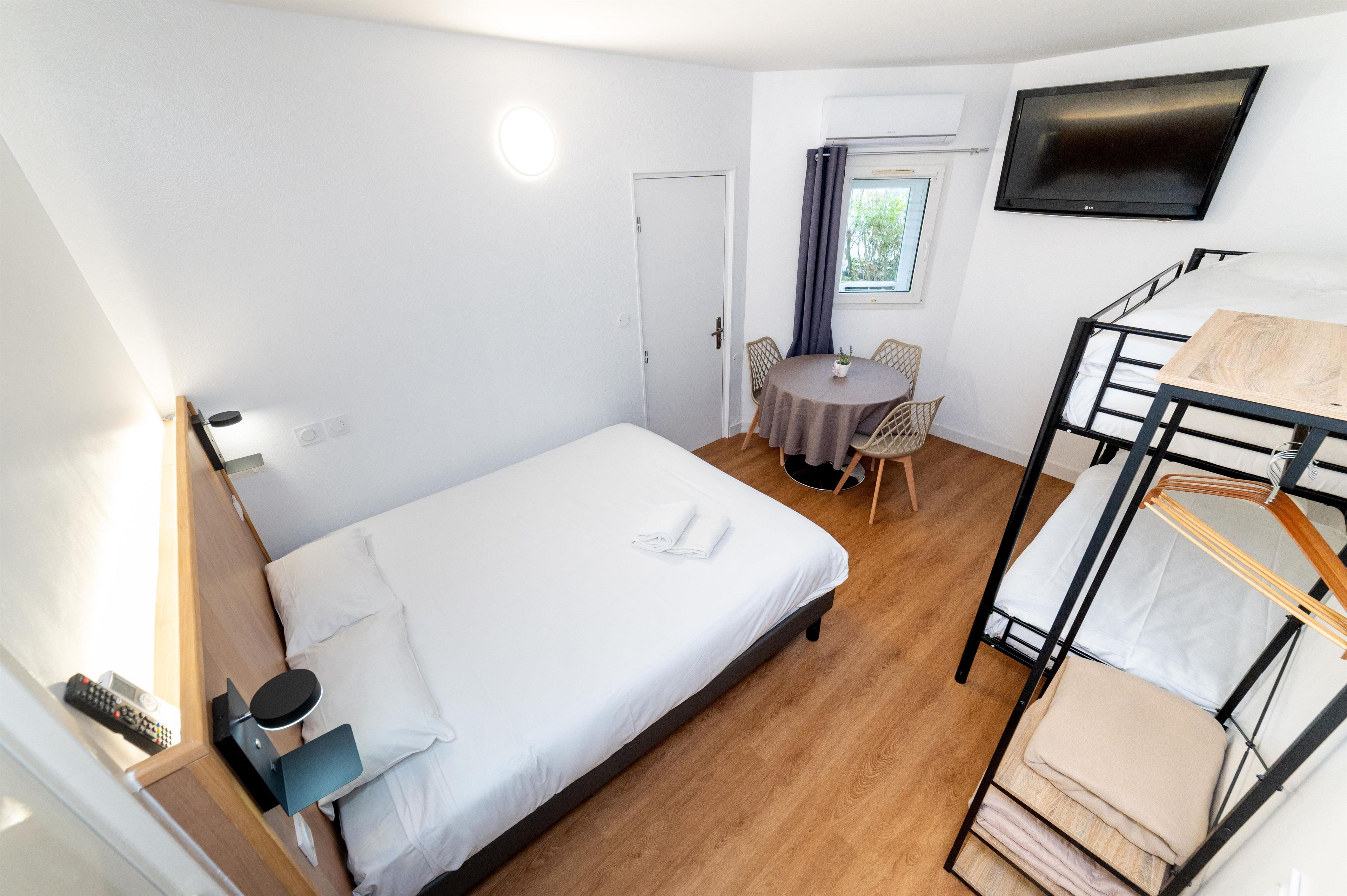 A room with a bunk bed, a single bed, a small table with chairs, a TV, and a window with curtains.