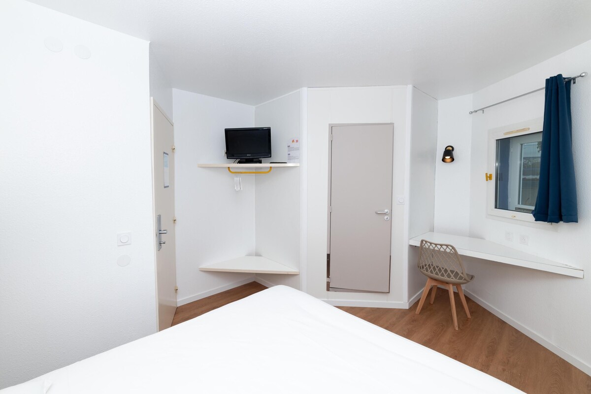 Una habitación con una cama, televisión en un estante, una puerta, una ventana con cortinas y una silla.