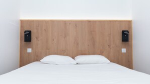 Un lit avec une literie blanche, une tête de lit en bois et deux haut-parleurs noirs fixés au mur.