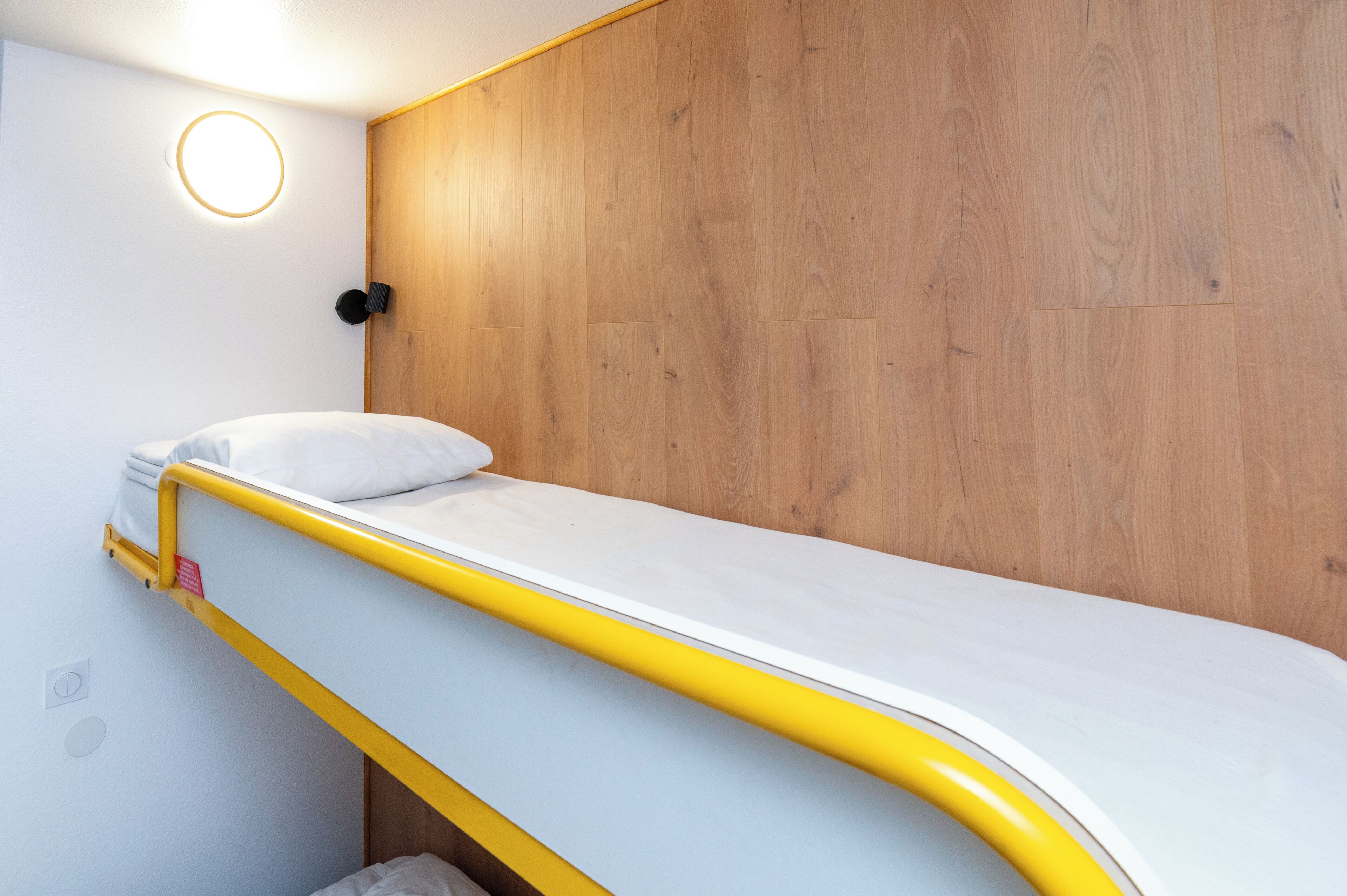 A bunk bed with a yellow frame and white mattress, a wooden panel wall, and a circular light fixture above the bed.