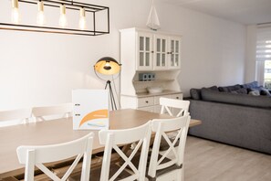 Un comedor con mesa de madera y sillas blancas, un armario blanco con puertas de cristal y un sofá gris al fondo.