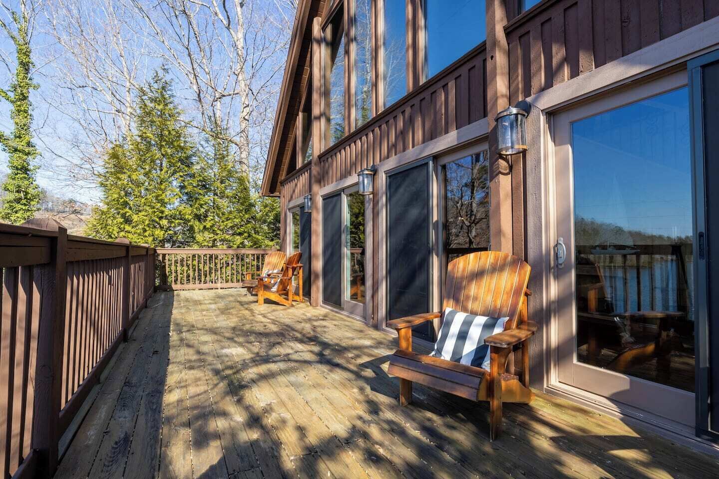 Eine Holzterrasse mit zwei Adirondack-Stühlen und einer Bank, großen Schiebeglastüren und Blick auf Bäume und ein Gewässer.