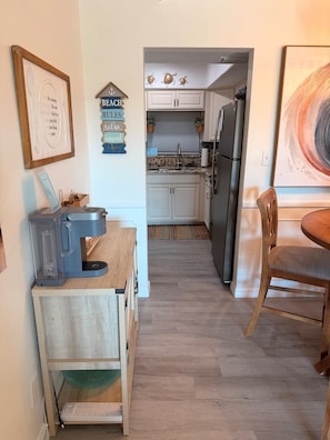 A kitchen with white cabinets, a coffee maker, and a dining area with a wooden table and chair. - Cozy 1 Bedroom by the Skyway Marina District (St. Petersburg)