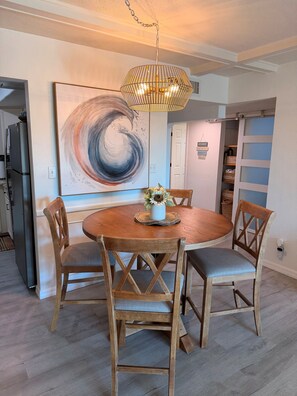 A dining area with a round wooden table, four chairs, a hanging light fixture, and a large abstract painting on the wall.