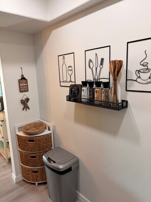 A kitchen area with a black metal shelf holding kitchen utensils, a trash bin, and wicker baskets.