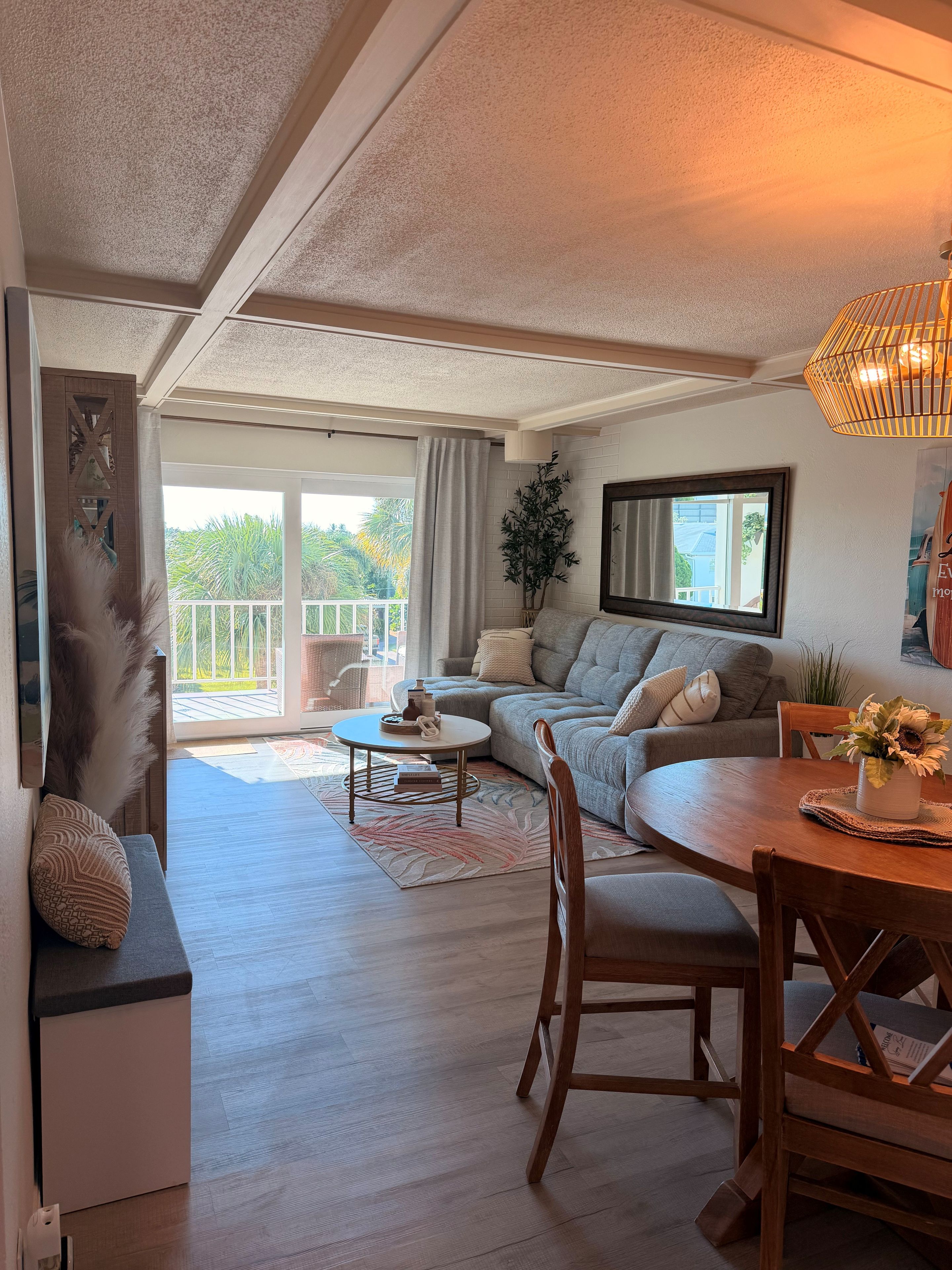 Un salon comprenant un canapé, une table à manger et une table basse. Une grande fenêtre offre une vue sur les palmiers.