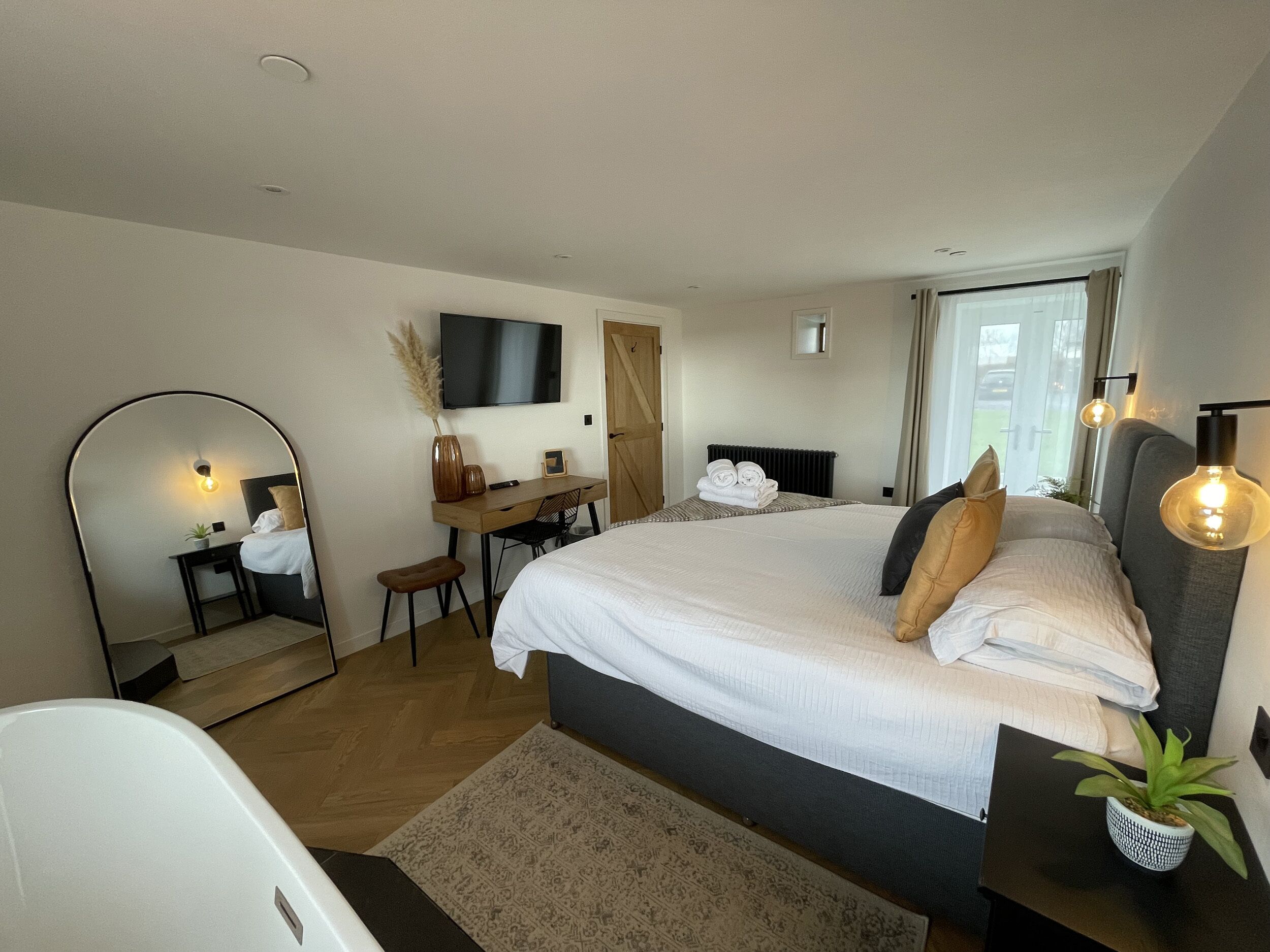 A modern bedroom with a large bed, a desk, a chair, a mirror, a TV, and a potted plant.