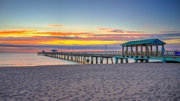 A beach with a wooden pier extending into the water, a covered pavilion, and a sunset in the background.
