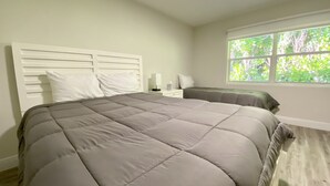 A bedroom with a large bed, a nightstand, a bedside table, and a window with a view of greenery.