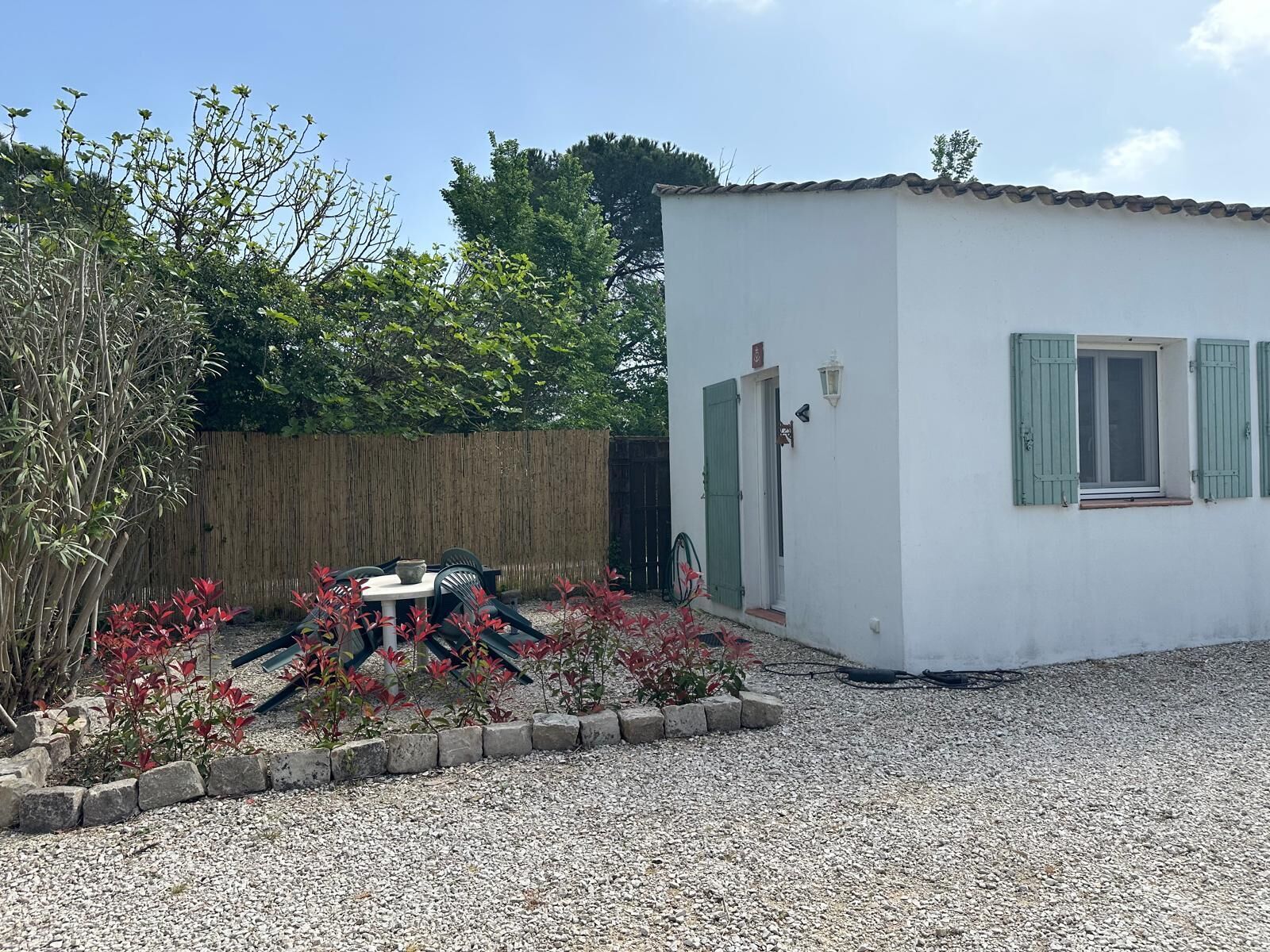 A small white building with a door and window, a wooden fence, and a gravel area with plants and a table.