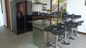 A modern kitchen with a large island, bar stools, a refrigerator, and a window with a view of rocks and greenery. - A luxury, modern and stylish sea view villa - 400 meters to the beach (Koh Phangan)