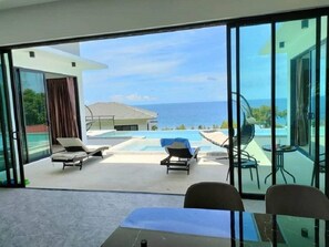 A modern room with a large sliding glass door leading to a pool area with lounge chairs and a view of the ocean. - A luxury, modern and stylish sea view villa - 400 meters to the beach (Koh Phangan)