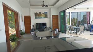 A modern living room with a flat-screen TV, a dining area with a glass table, and a large sliding glass door leading to a balcony. - A luxury, modern and stylish sea view villa - 400 meters to the beach (Koh Phangan)