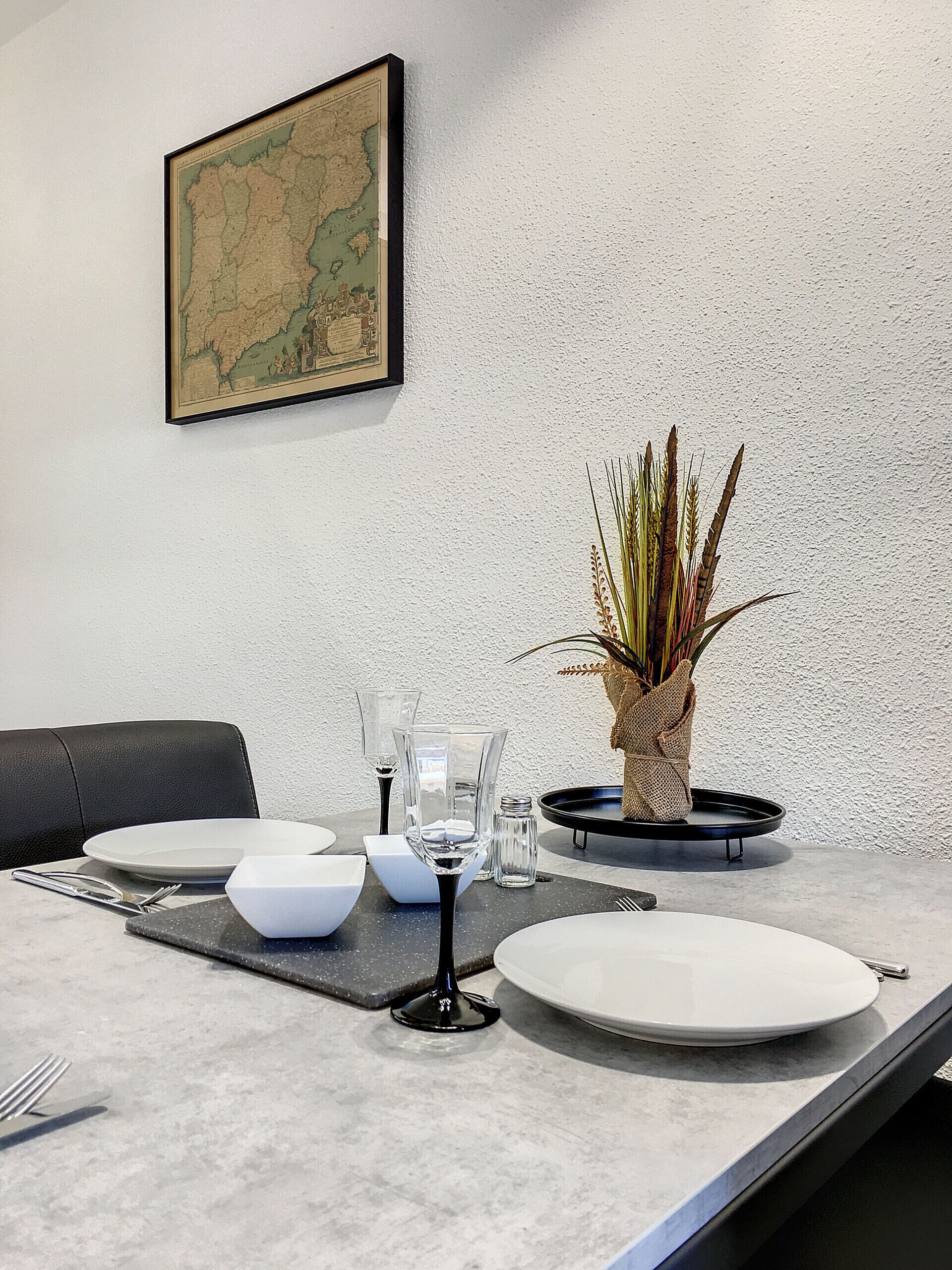 A dining table set with plates, glasses, and cutlery, a vase with plants, and a framed map on the wall.