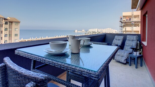 Un balcon doté de meubles en osier, d’une table et offrant une vue sur la mer et le paysage urbain.