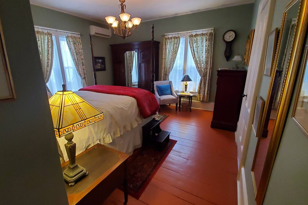 A room with a bed, a chair, a table, a lamp, a clock, and a mirror.