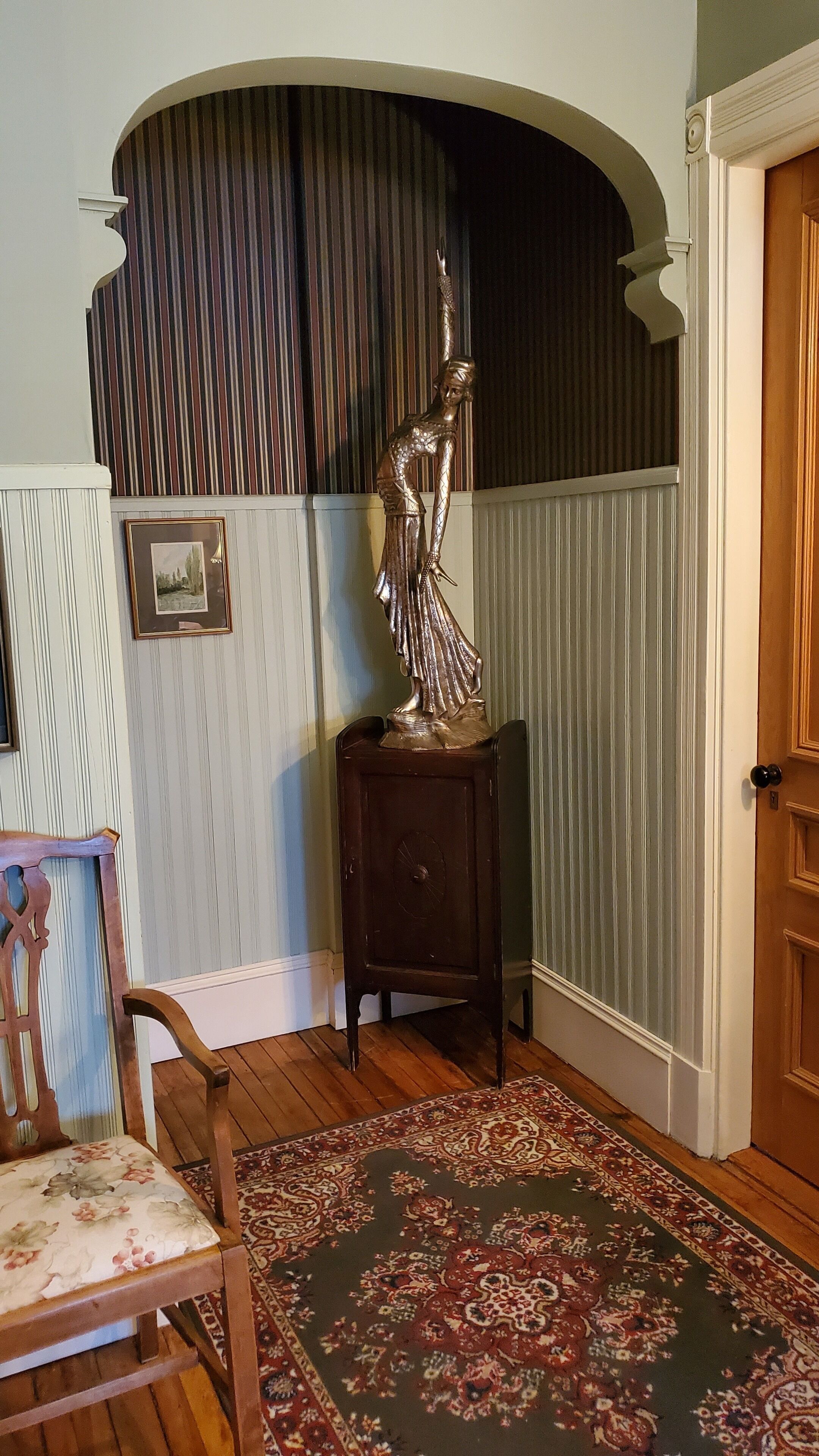 A room with a wooden chair, a dark wooden side table with a statue, a patterned rug, a door, and a striped wall with a picture frame.