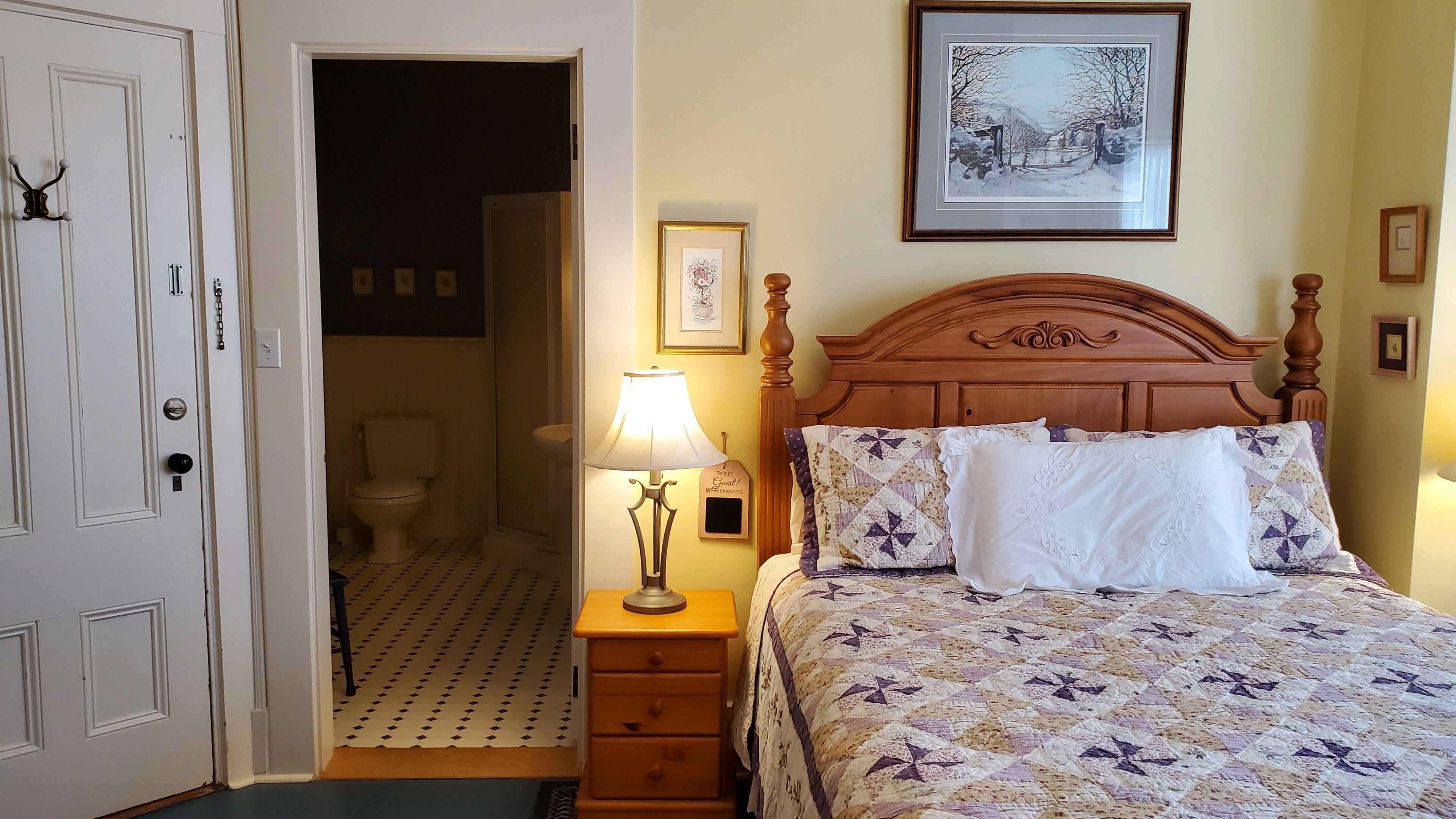A hotel room with a wooden bed, a bedside lamp, a framed picture on the wall, and a door leading to a bathroom.