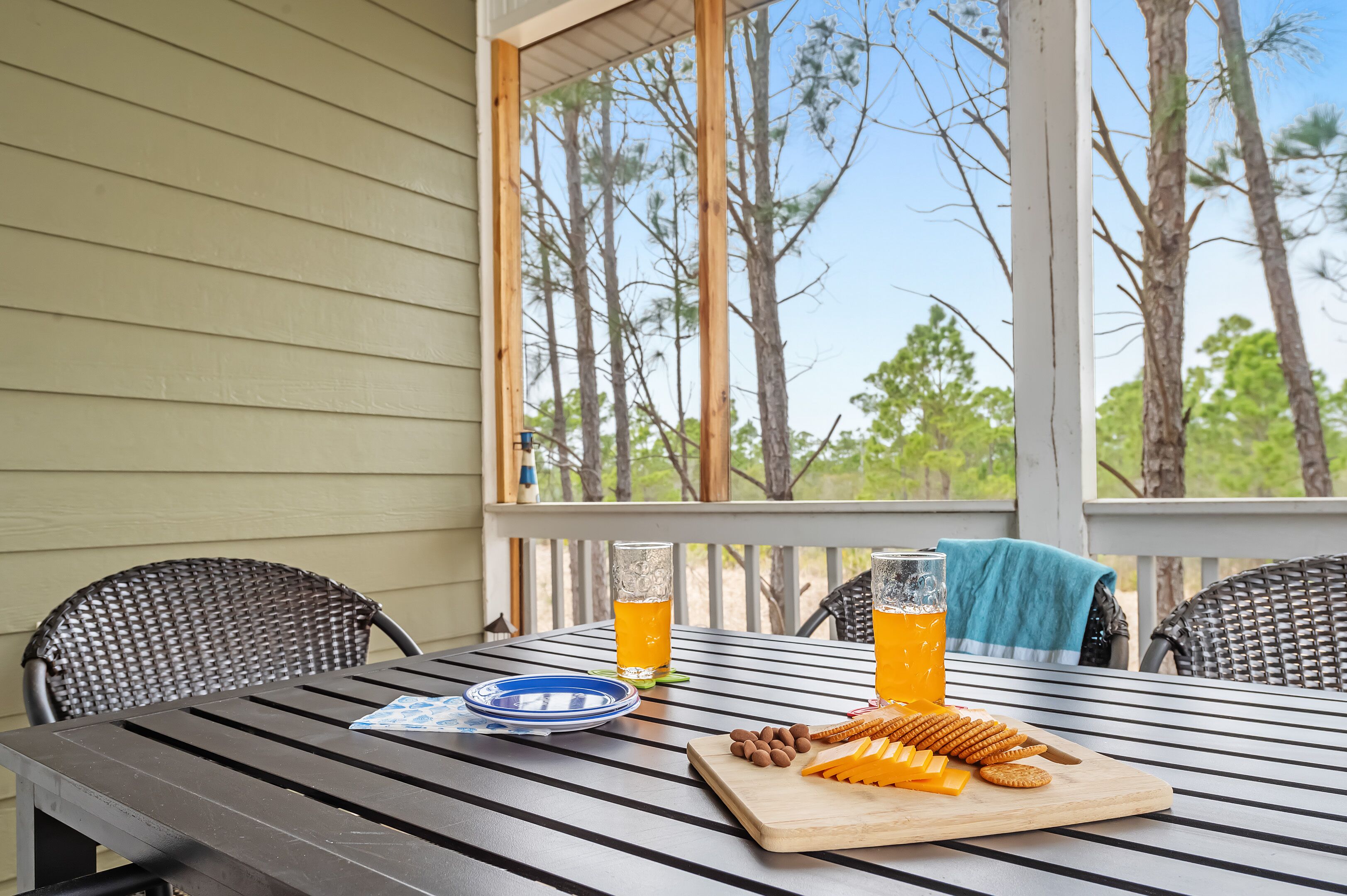 Une table à manger donnant sur des arbres, visibles à travers une fenêtre, une assiette, deux verres de jus d’orange et une planche à découper avec du fromage et des crackers.