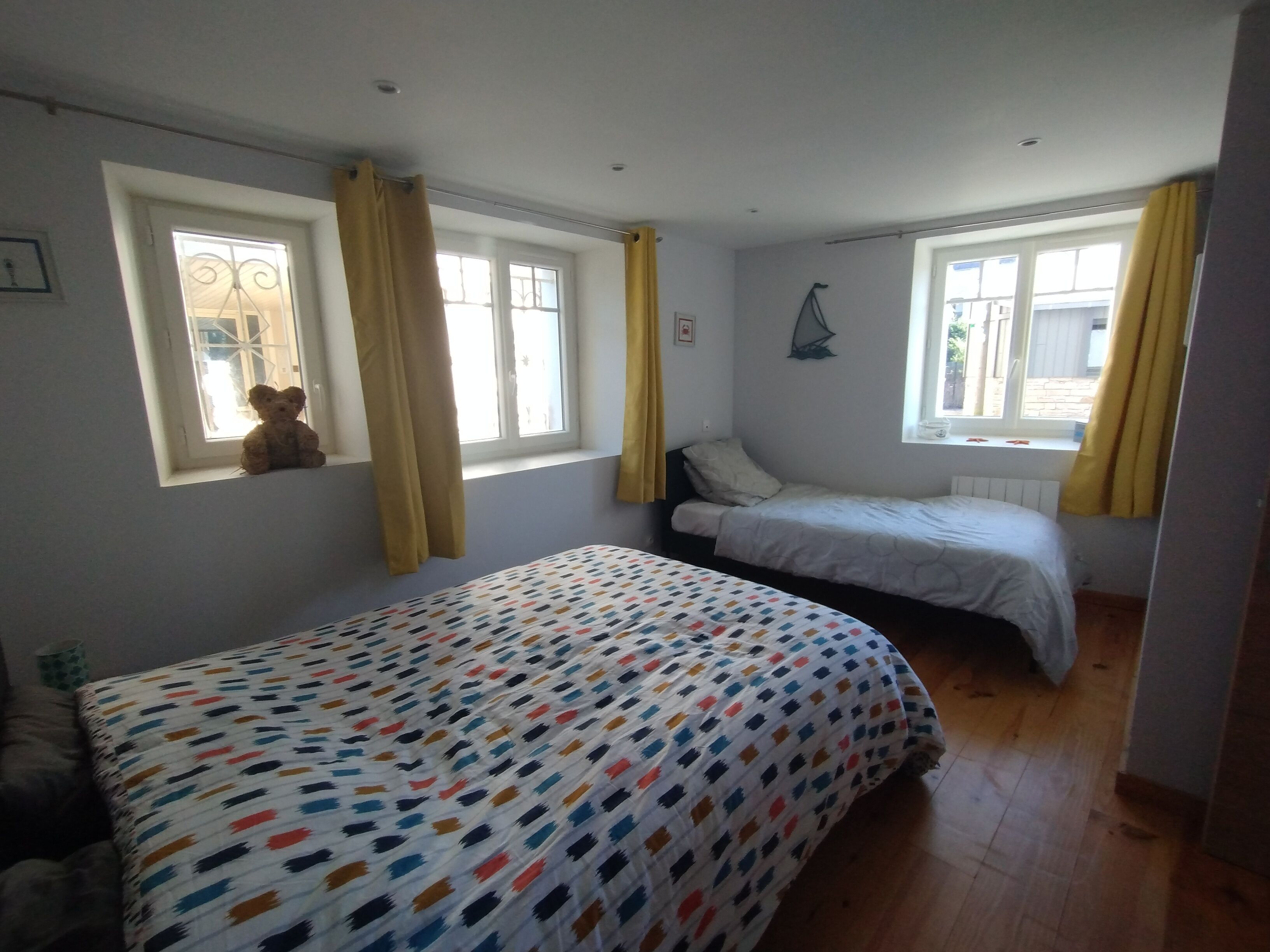 Een slaapkamer met twee bedden, een raam met gordijnen, een teddybeer op de vensterbank, en een wandlamp.