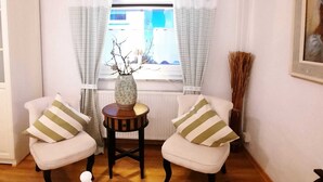 Ein Zimmer mit zwei Stühlen, einem kleinen Tisch, einer Vase und einem Fenster mit Vorhängen.