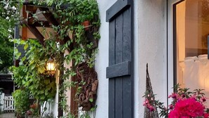 Ein weißes Gebäude mit einem dunkelholzfarbenen Fensterladen, einer hängenden Pflanze mit grünen Blättern und einer Kübelpflanze mit rosa Blüten.