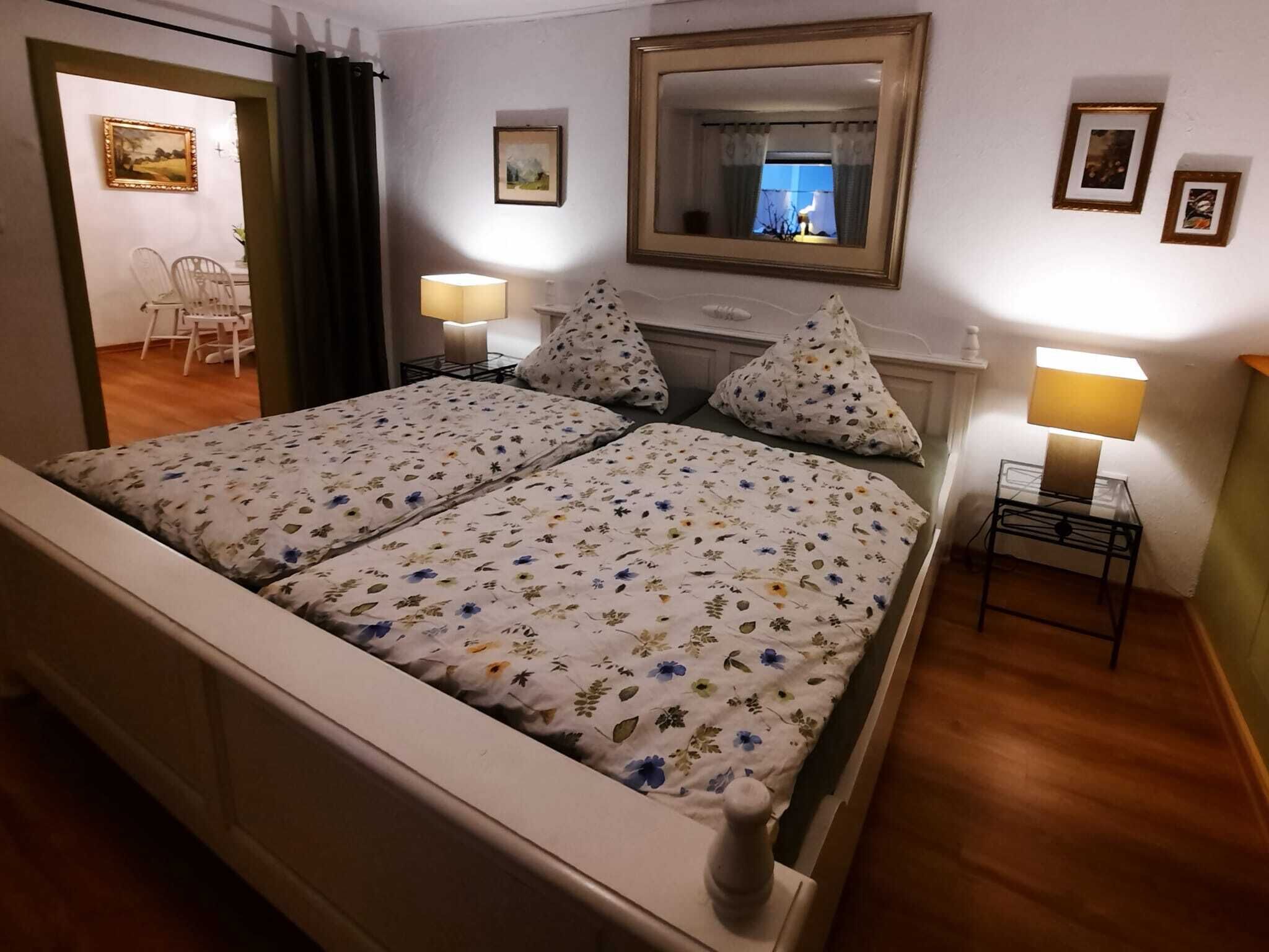 A double bed with floral bedding, a bedside table with a lamp, a mirror above the bed, and a small table with a lamp in a hotel room.