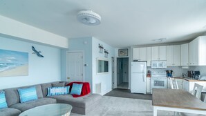 A modern living room with a grey sofa, a round coffee table, and a dining area with a wooden table and chairs. A kitchen with white cabinets and appliances is visible in the background. - Beach, beach, in historic Cape Charles. Lovely 2BR Apt. 1 Block from beach (Cape Charles)