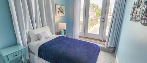 A bedroom with a bed, a nightstand, a lamp, a window with curtains, and a view of the outside.
