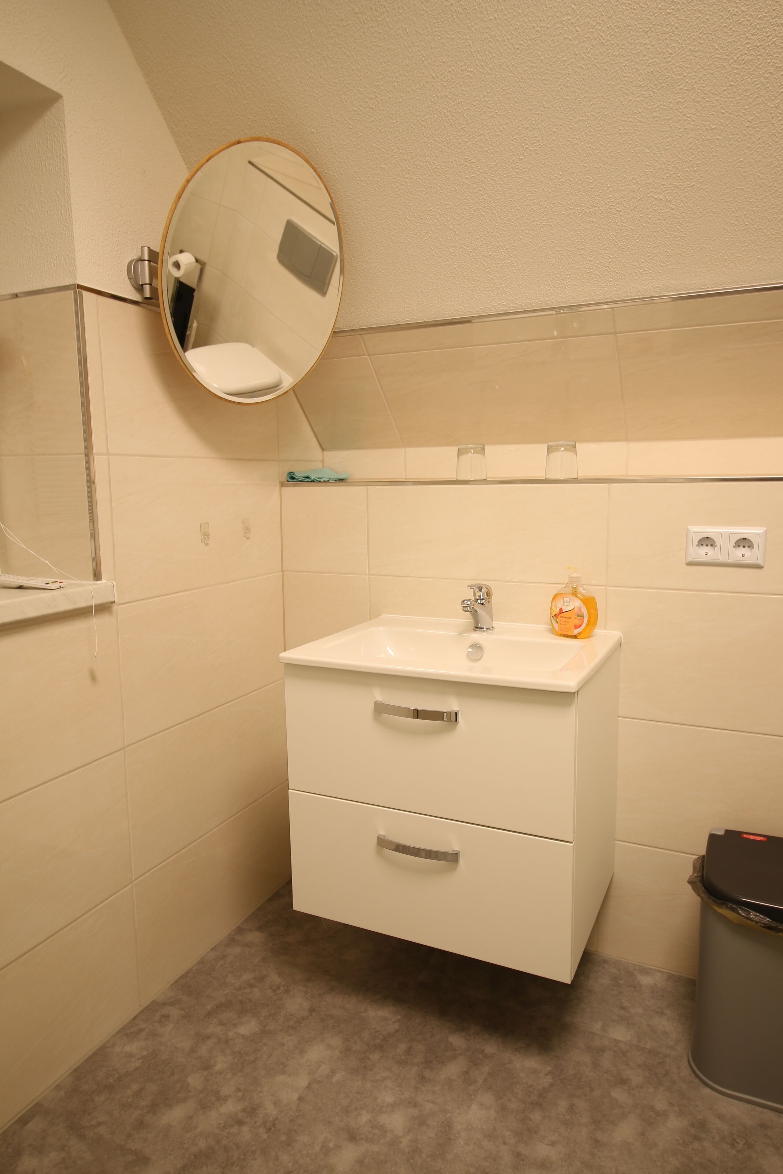 Een badkamer met een witte wastafel, een spiegel, een prullenbak en een handdoekhouder.
