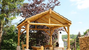 A wooden gazebo with a table and benches, surrounded by trees and a stone wall. - Vacation apartment in a half-timbered house (Jonsdorf)