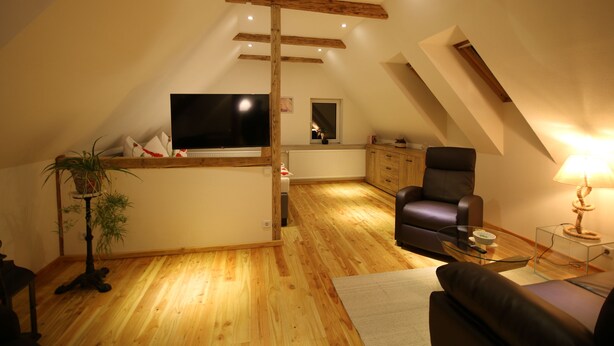 A loft-style living space with a high ceiling, wooden beams, a flat-screen TV, a sofa, a chair, and a lamp. - Vacation apartment in a half-timbered house (Jonsdorf)