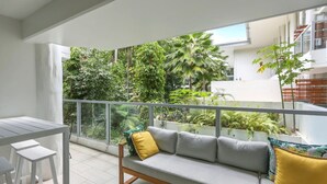 A balcony with a grey sofa, white stools, and a glass railing overlooking a lush garden with various plants and a palm tree.