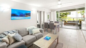 A modern living room with a grey sofa, a coffee table, and a painting on the wall. There is a dining area with a table and chairs, and a balcony with a sofa and plants.