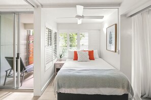 Een slaapkamer met een bed, witte muren, een plafondventilator en een raam met jaloezieën.