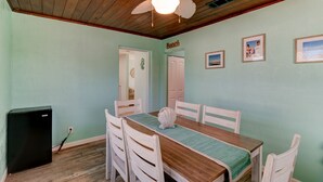 Un comedor con mesa de madera, sillas blancas, ventilador de techo y refrigerador.