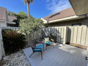 A wooden deck with two wicker chairs and a small table, surrounded by a white fence and greenery.