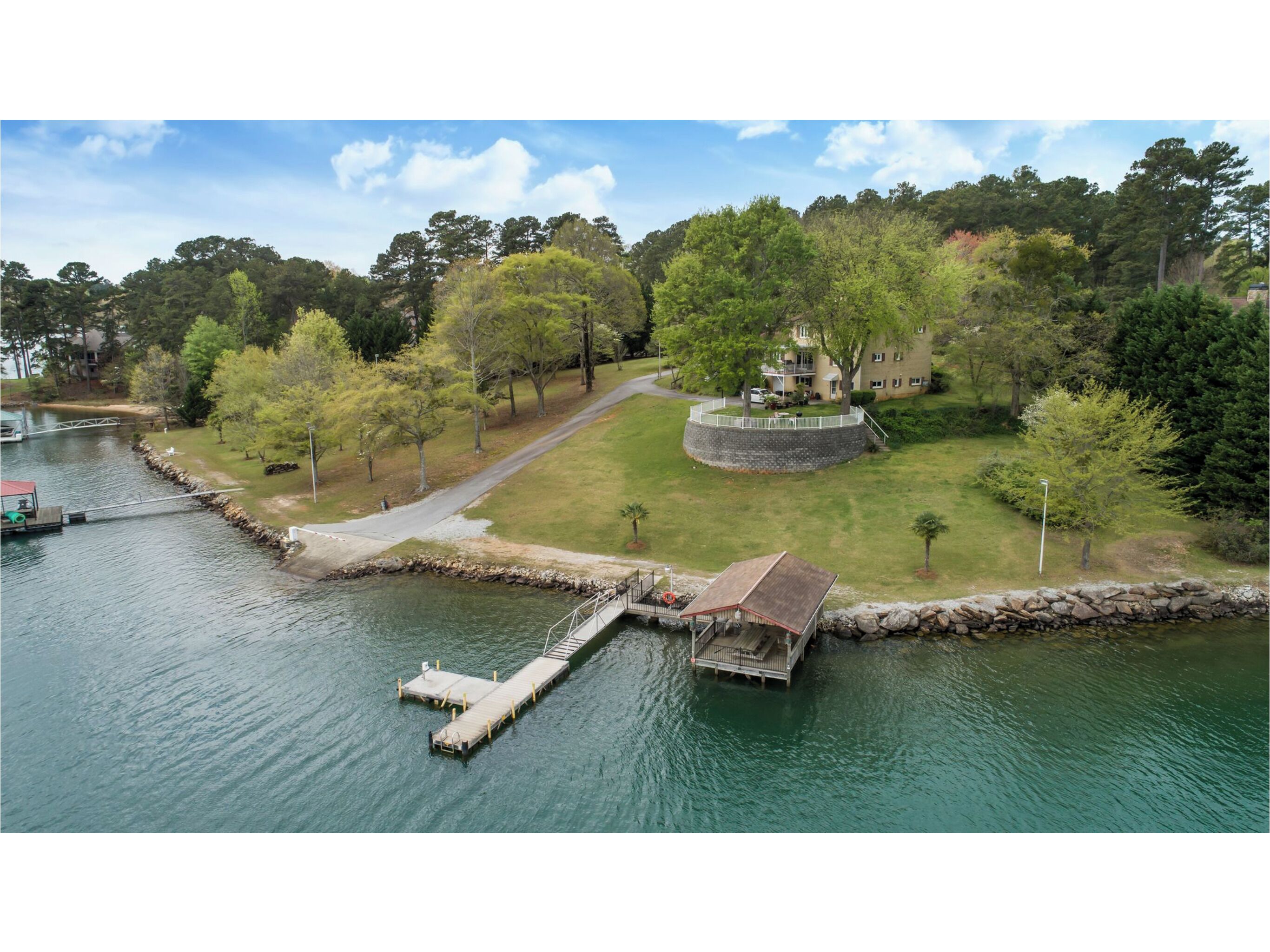 A riverside park with a wooden dock, a stone bridge, and a building surrounded by trees.