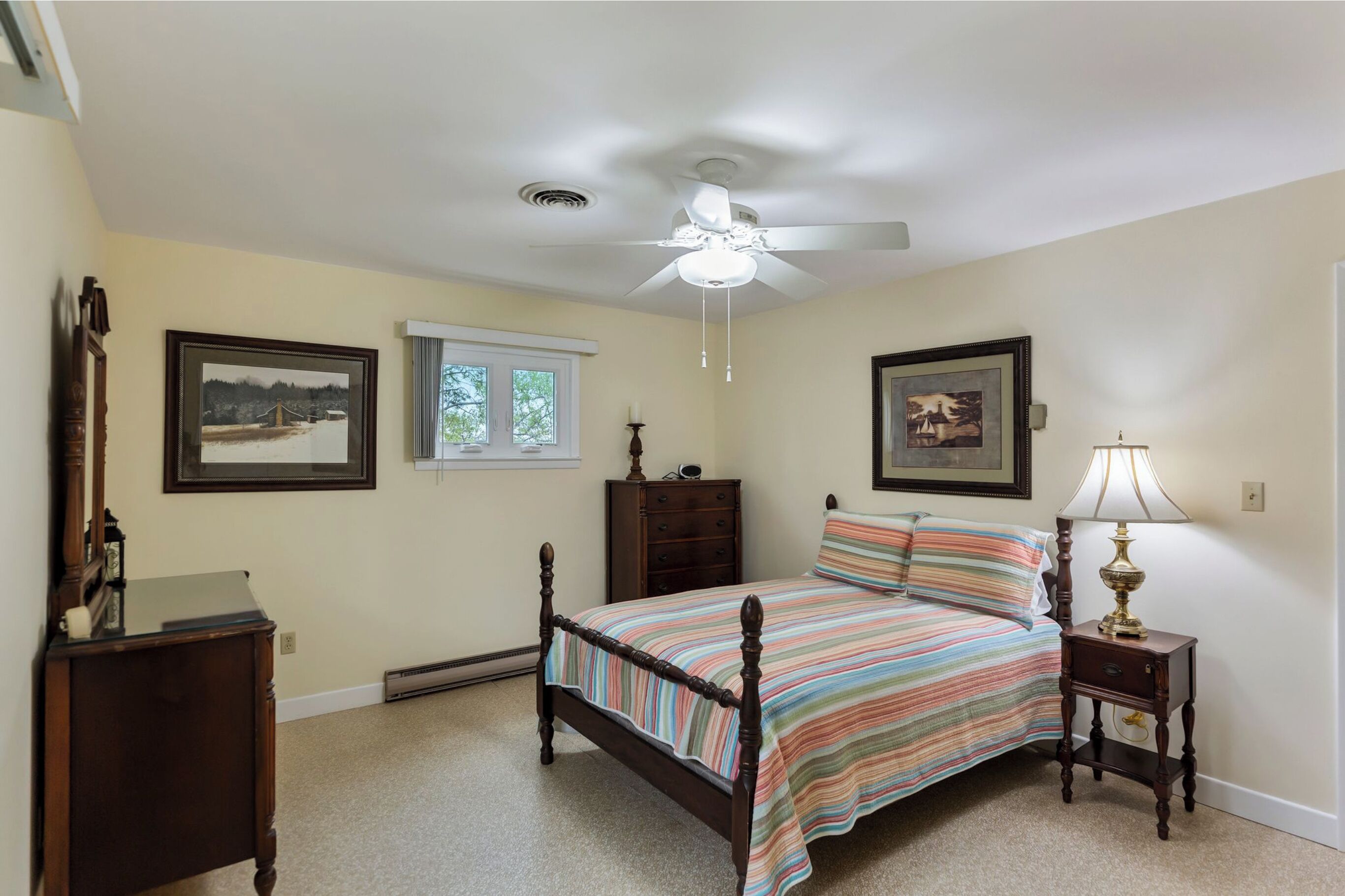 Une chambre à coucher comprenant un lit, un ventilateur de plafond, une table de chevet avec une lampe, une commode et deux tableaux encadrés accrochés au mur.