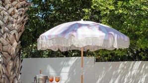 Ein pink-weiß gestreifter Schirm mit Fransen, ein pinkfarbener Tisch mit Champagnerkühlbag und zwei Gläsern sowie eine weiße Wand mit Grünpflanzen im Hintergrund.