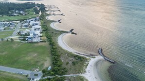 An aerial view of a coastal area with a long stretch of beach, a road curving along the coast, and a row of houses adjacent to the beach. - Gorgeous 4BR Home With Panoramic Sunsets & Walking Distance to Private Beach! (Cape Charles)