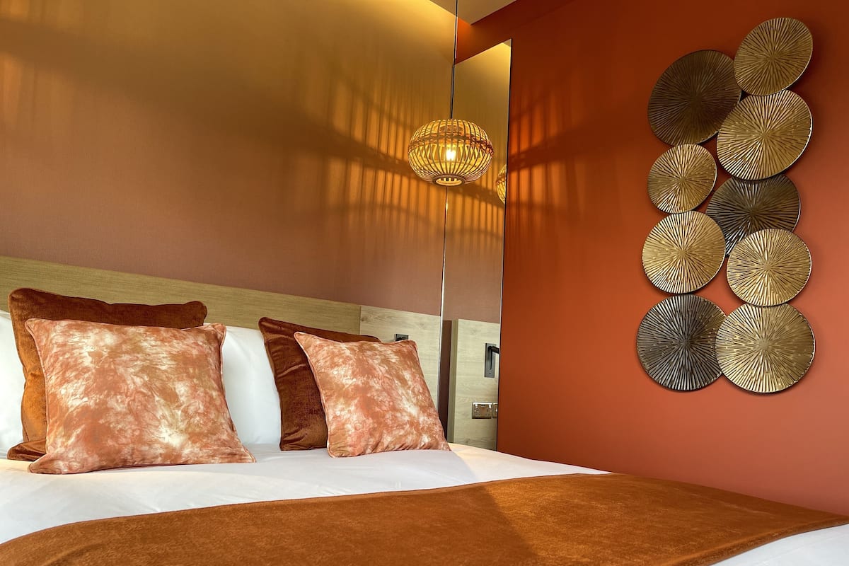 A modern hotel room with a large bed, multiple pillows, a decorative wall piece, and a hanging light fixture.