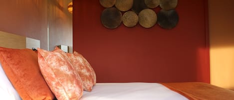 A neatly made bed with white and orange bedding, a red wall with circular wall art, and a hanging light fixture.
